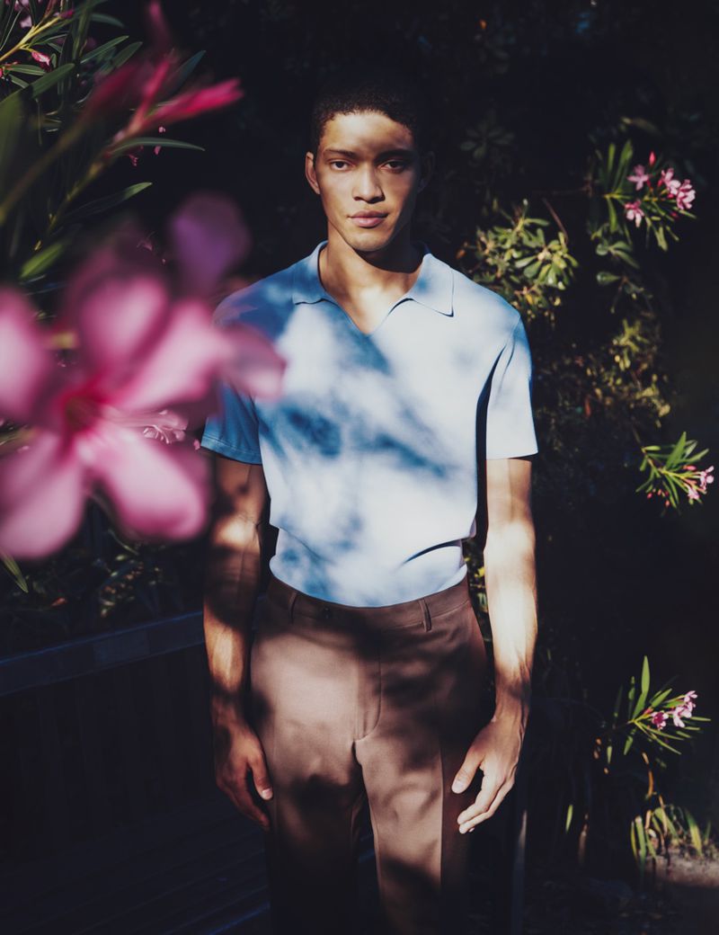 Model Jeranimo van Russel in a light blue polo and brown trousers standing amidst lush foliage with dappled sunlight.