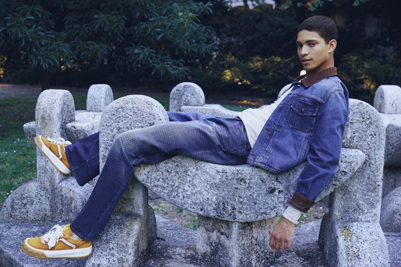 Model Jeranimo van Russel lounging on a carved stone bench wearing a blue denim jacket, matching jeans, and yellow sneakers.