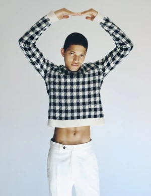 Model Jeranimo van Russel wearing a cropped black and white check sweater and white trousers with arms raised against a white backdrop.