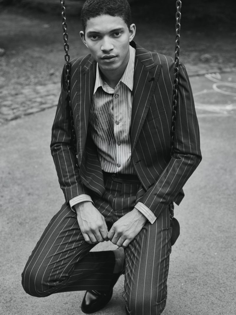 Model Jeranimo van Russel wearing a pinstripe suit and striped shirt while crouched on a chain swing outdoors in black and white.
