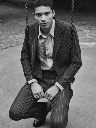 Model Jeranimo van Russel wearing a pinstripe suit and striped shirt while crouched on a chain swing outdoors in black and white.