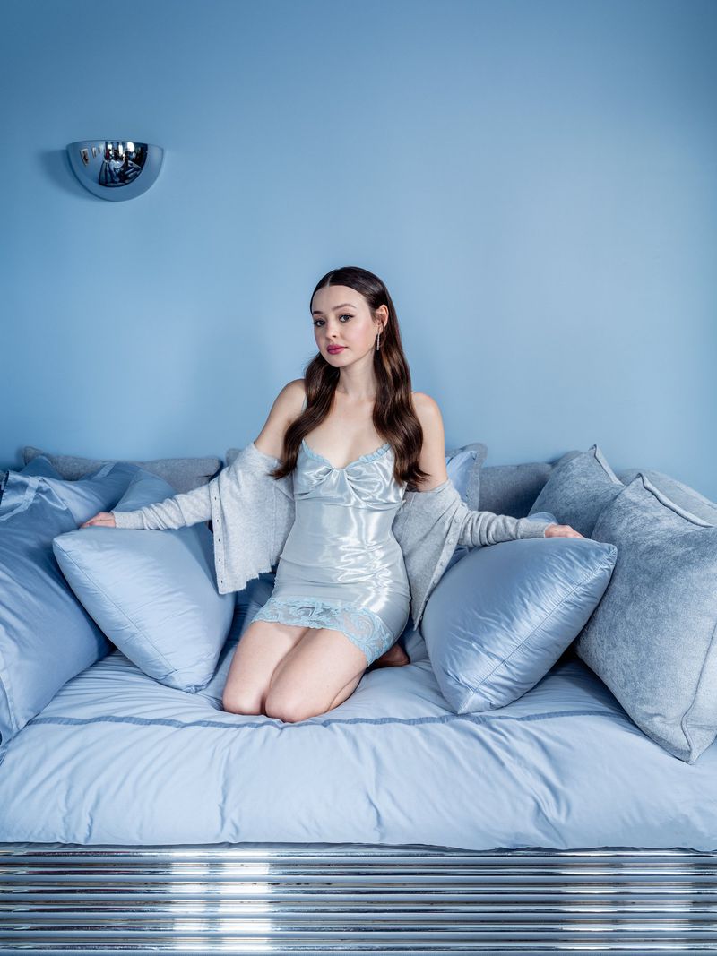 Eileen Kelly kneeling on a light blue daybed with matching satin pillows against a monochrome blue wall.