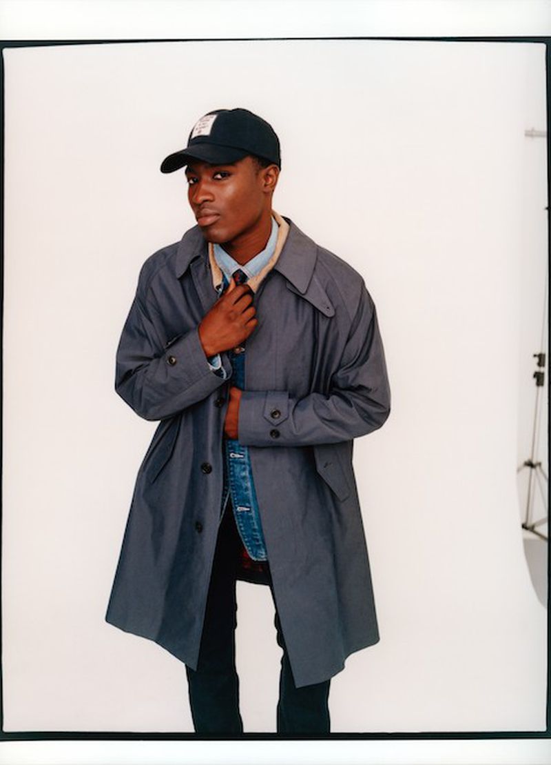 Model Momo Ciss wearing a grey overcoat, denim jacket, and black baseball cap against a white backdrop.