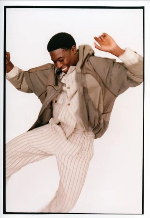 Model Momo Ciss jumping mid-air in a cream pinstripe outfit and hooded jacket against a white studio background.