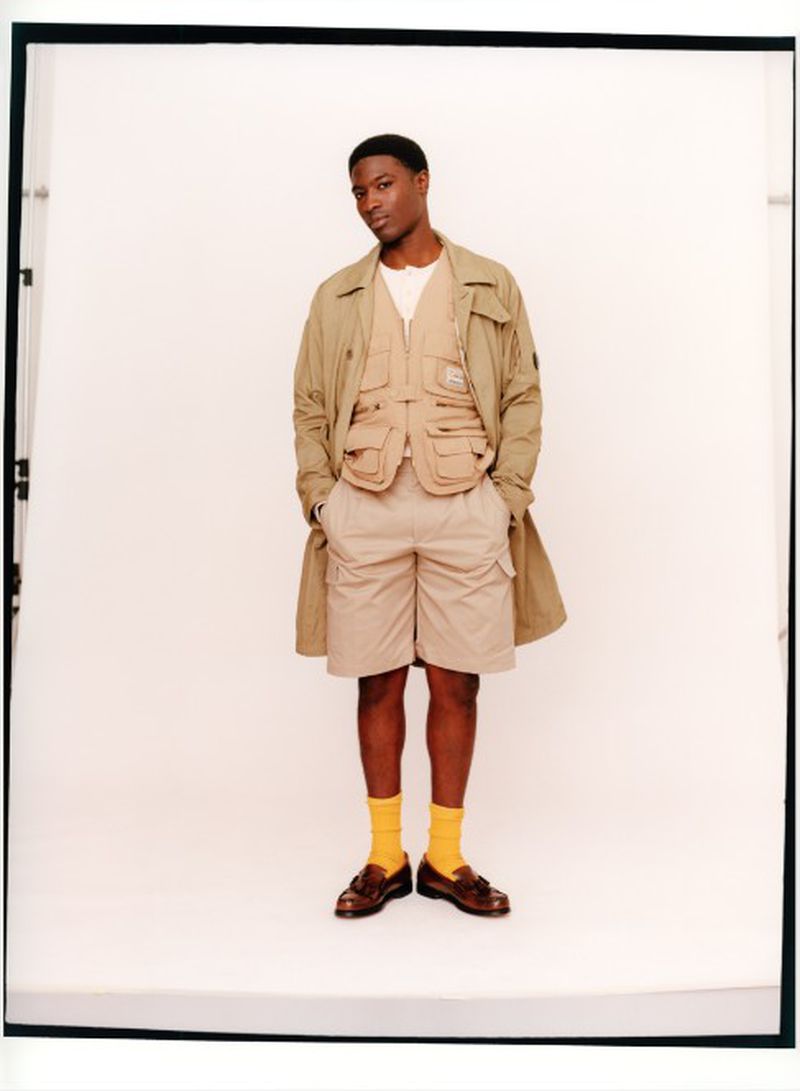 Model Momo Ciss wearing a beige utility vest, matching shorts, long coat, and yellow socks in a studio portrait.