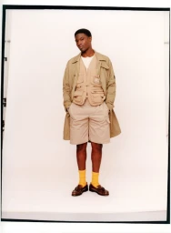 Model Momo Ciss wearing a beige utility vest, matching shorts, long coat, and yellow socks in a studio portrait.