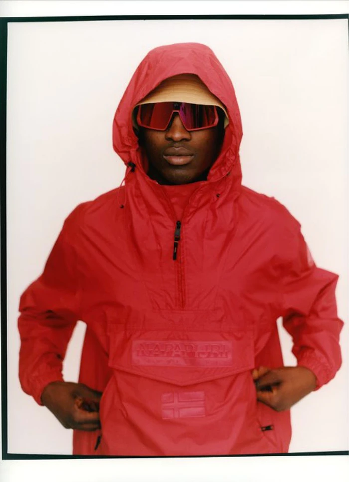 Model Momo Ciss wearing a red hooded windbreaker jacket and tinted shield sunglasses against a plain white backdrop.