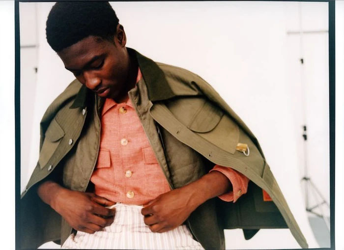 Momo Ciss wearing an olive utility jacket over a coral buttoned shirt and striped trousers against a white studio background.