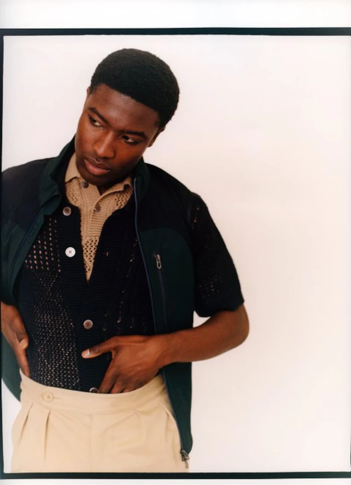 Model Momo Ciss wearing a layered black knit shirt, dark vest, and pleated beige trousers against a white backdrop.