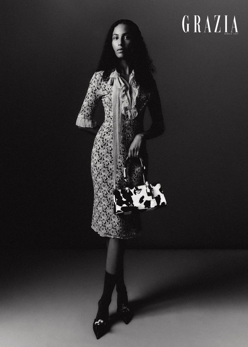 A full-length studio shot in black and white of an adult woman in a floral dress holding a cow-print purse, complemented by black heels.