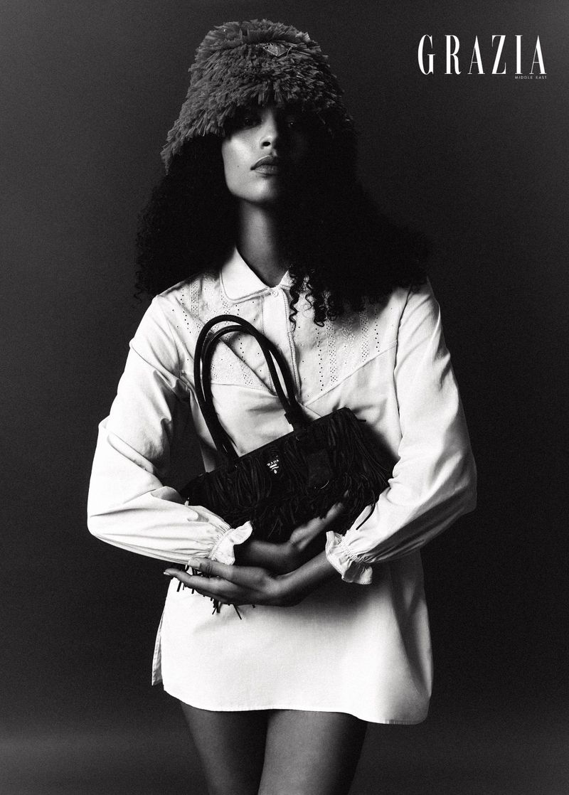 Black and white portrait of a woman wearing a fur hat and holding a fringed bag, showcasing fashion and editorial style in a studio setting.