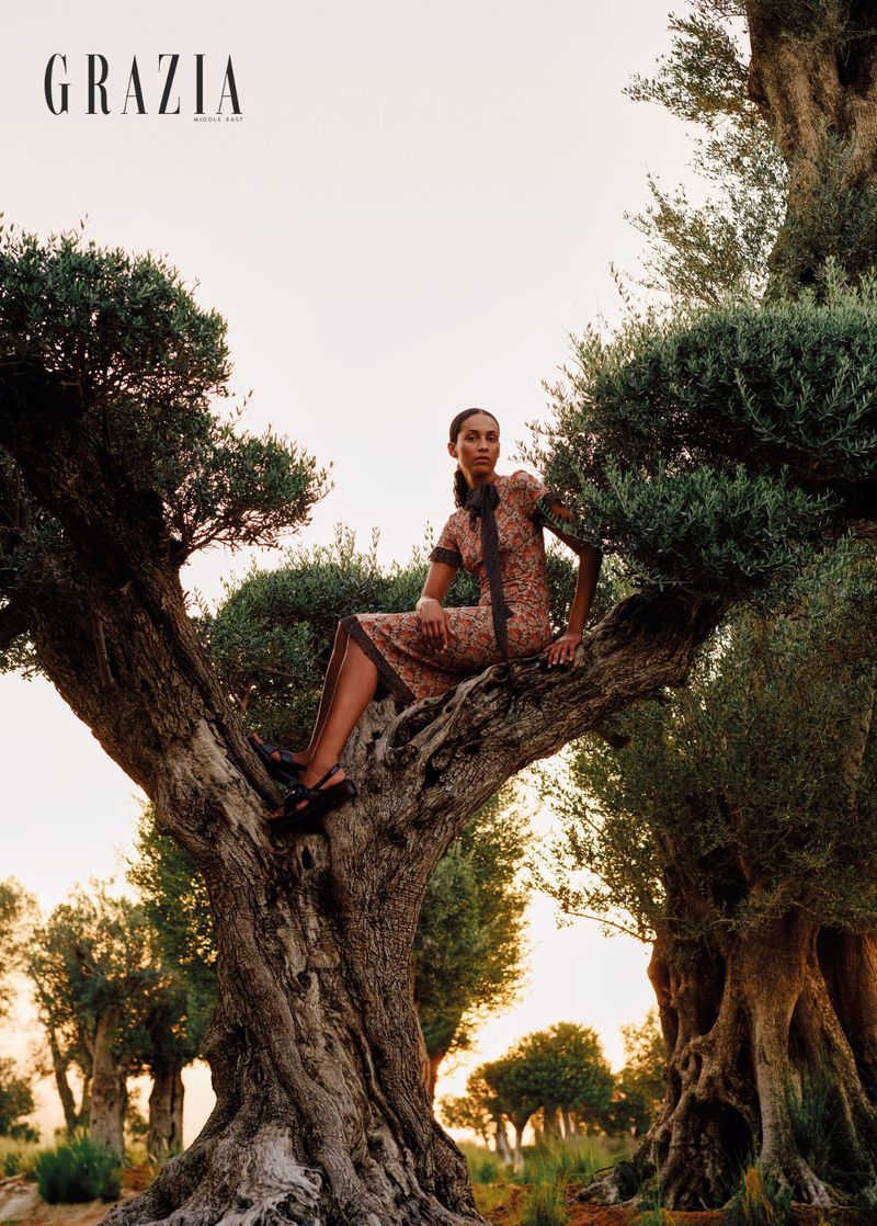 Fashion editorial shot of a woman in a patterned dress sitting in a large, old tree in a sunlit grove. 'Grazia' magazine logo visible.