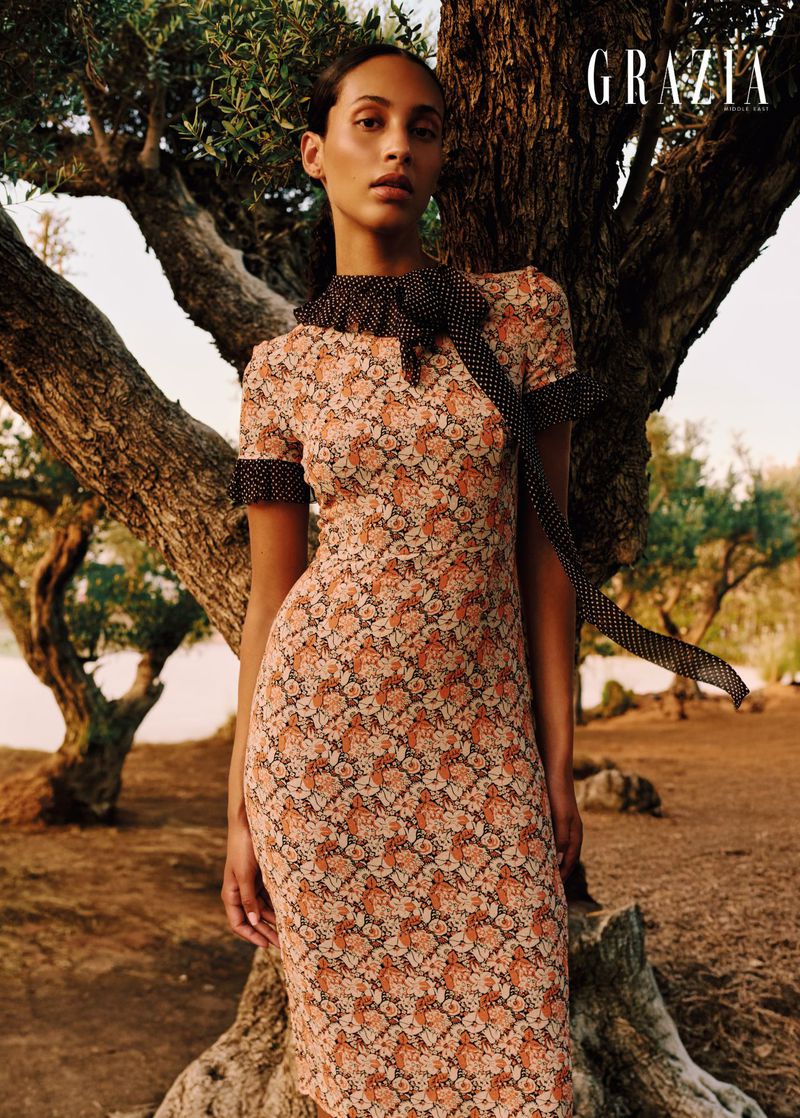 Model Anissa Bakhti poses in a floral dress with a polka dot scarf beside a large olive tree.
