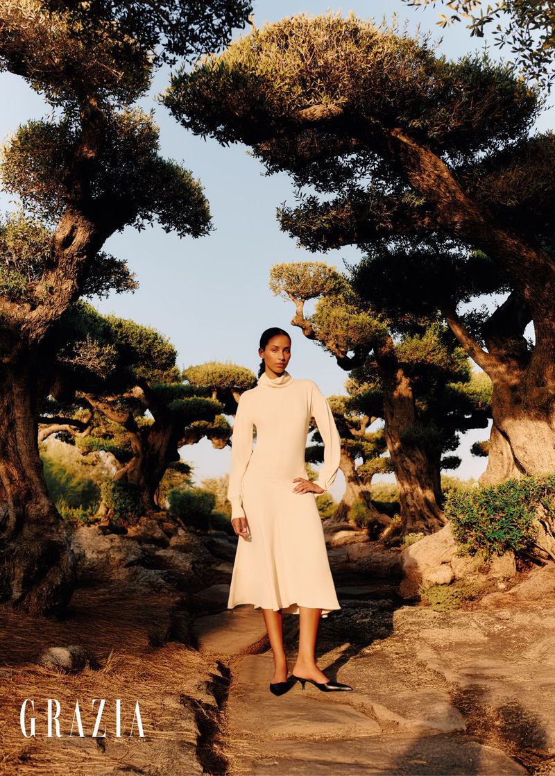 An adult woman poses in a cream-colored dress and low heels amid topiary trees for a fashion editorial, illuminated by natural light.
