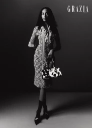 Model Anissa Bakhti posing in a patterned midi dress with a ruffled neckline and holding a printed handbag in black and white.