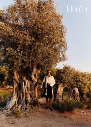 Anissa Bakhti stands in a white blouse and black button-front skirt beside an ancient gnarled olive tree.