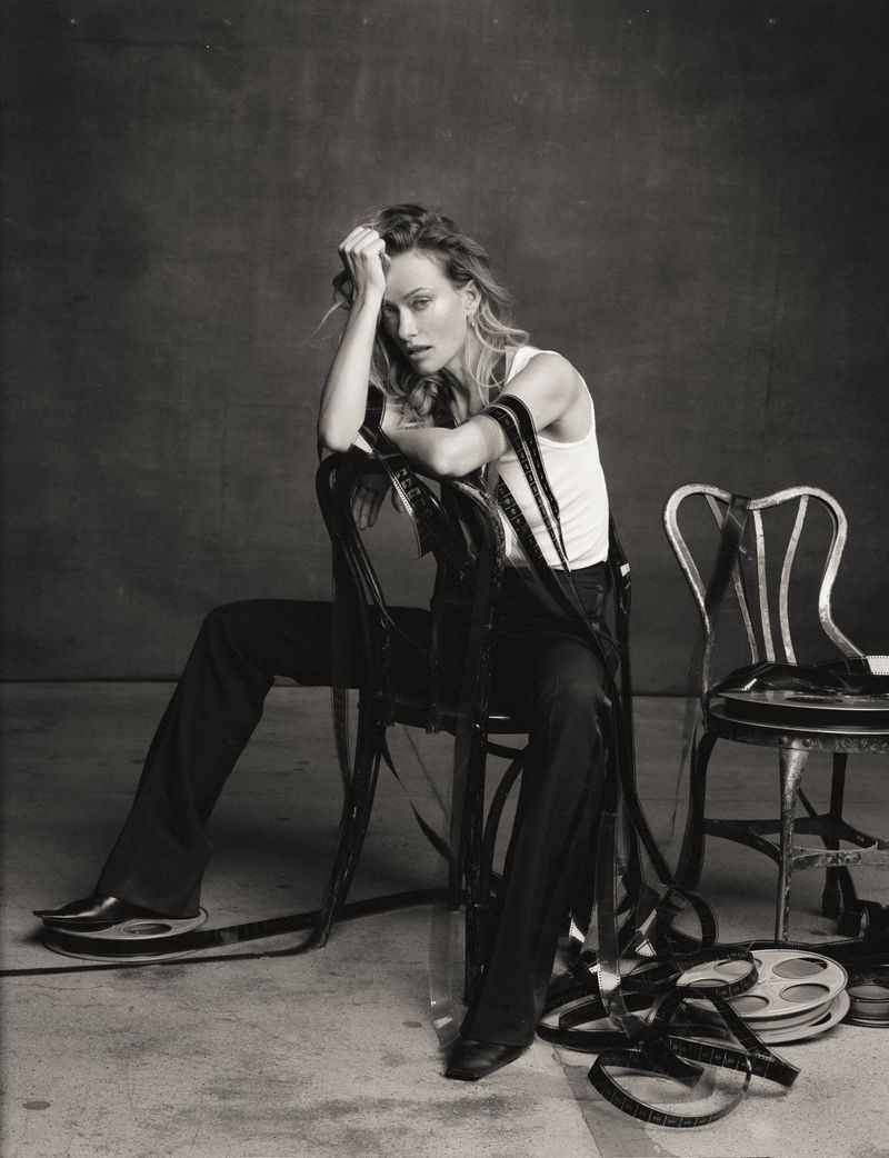 Olivia Wilde seated backwards on a chair draped in tangled film strips wearing a white tank top and black trousers in black and white.
