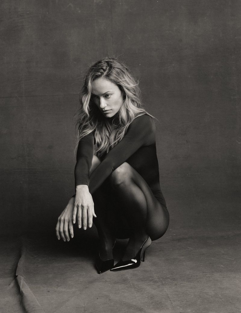 Olivia Wilde crouching in a long-sleeve black bodysuit, sheer tights, and patent high heels against a textured backdrop.