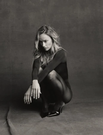 Olivia Wilde crouching in a long-sleeve black bodysuit, sheer tights, and patent high heels against a textured backdrop.