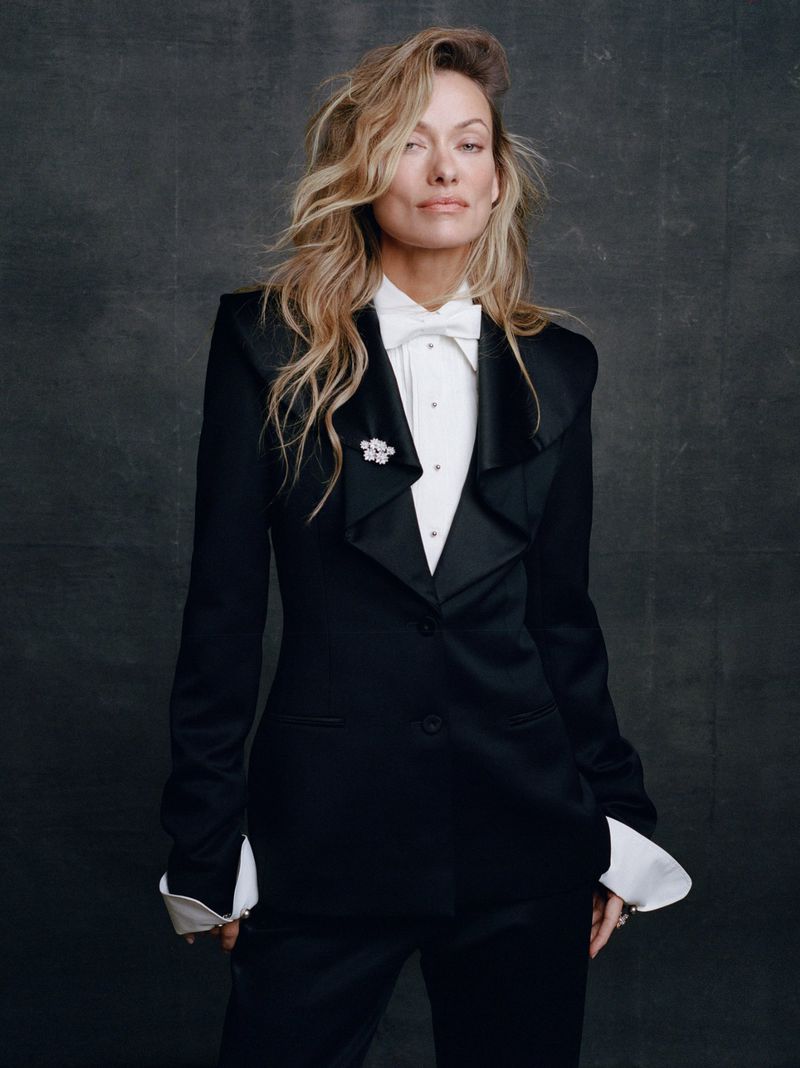 Olivia Wilde wearing a tailored black tuxedo suit with a white shirt, white bow tie, and sparkling brooch against a dark backdrop.