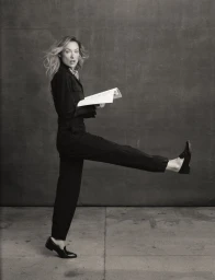Olivia Wilde kicking her leg up while holding an open script against a textured backdrop in a black jumpsuit.
