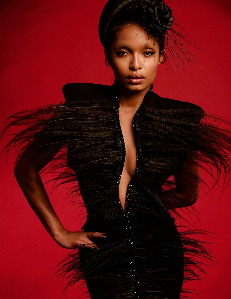 Yara Shahidi posing in a structured black feathered dress with a matching floral fascinator against a rich red background.