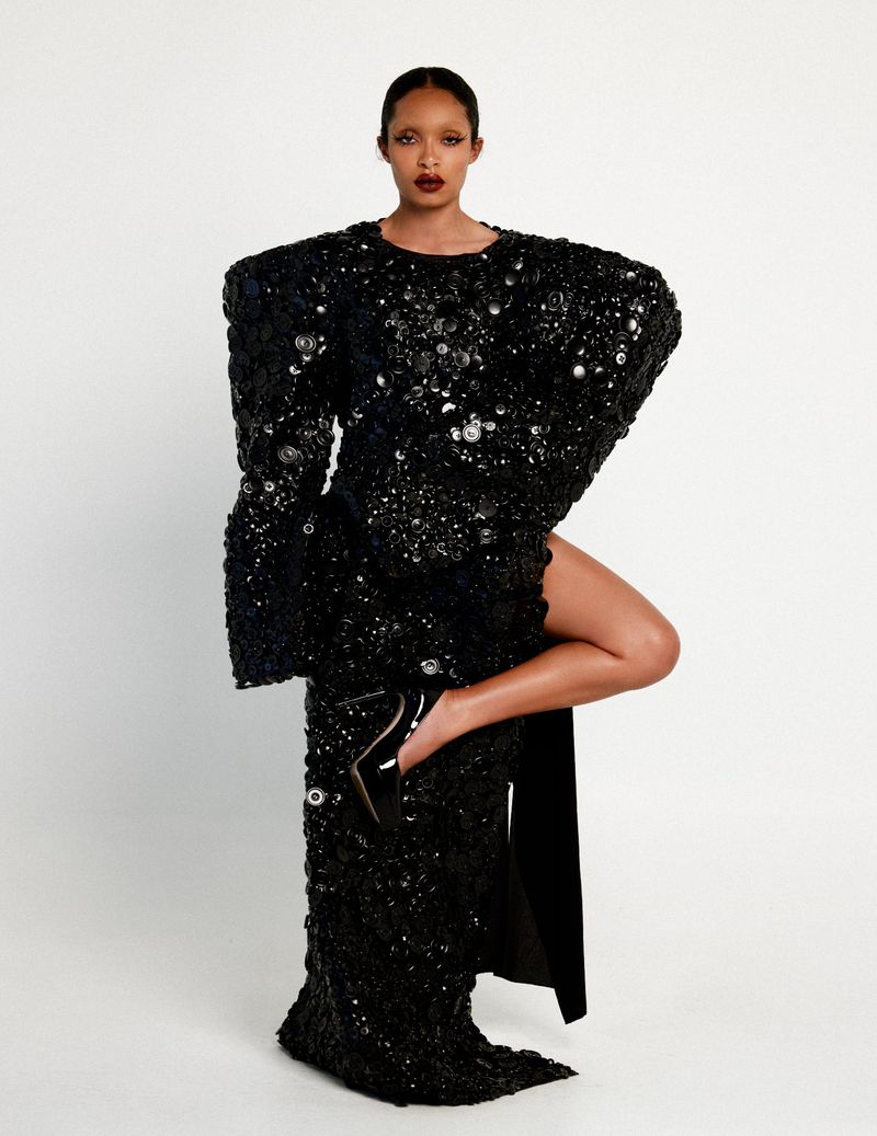 Yara Shahidi posing in an exaggerated structural black sequin gown with a high leg slit against a clean white seamless backdrop.