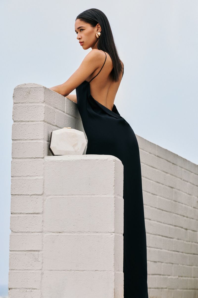 A woman in a backless black gown leans on a white brick architectural wall next to a geometric clutch against a pale sky.