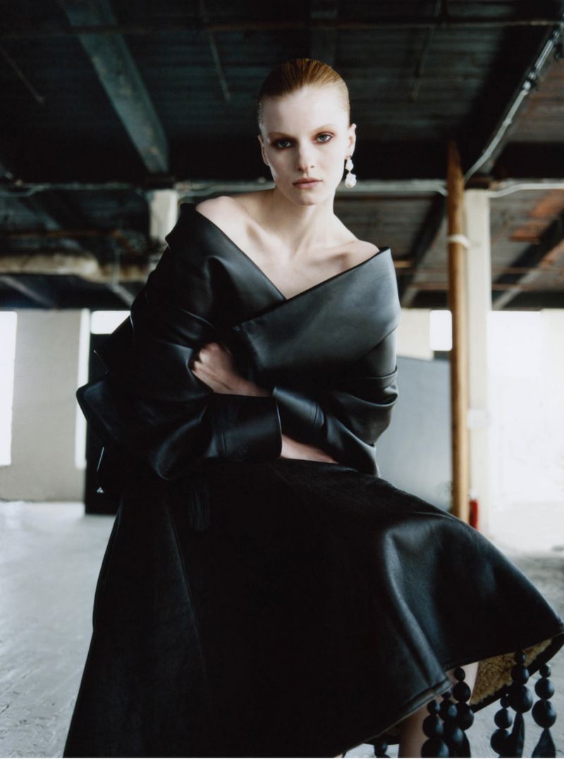 Model Lainey Hearn wearing a sculptural off-the-shoulder black leather dress in an industrial loft space.