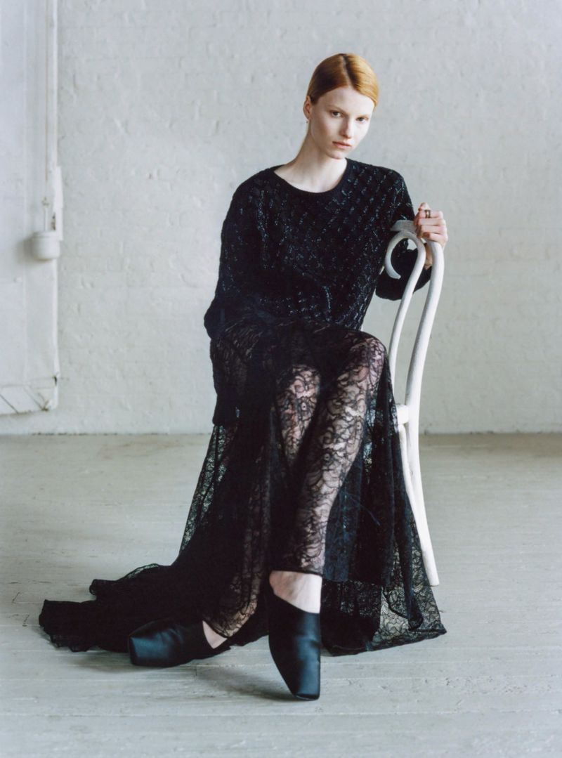 Model Lainey Hearn seated on a white bentwood chair wearing a black textured knit top and black floral lace skirt in a studio.