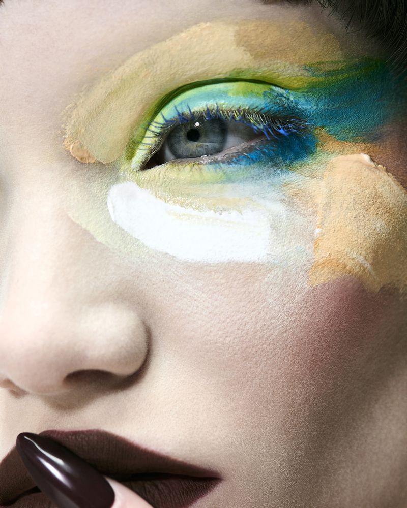 Macro shot of a model with artistic paint makeup around the eye in teal, green, and beige, paired with deep burgundy lipstick.