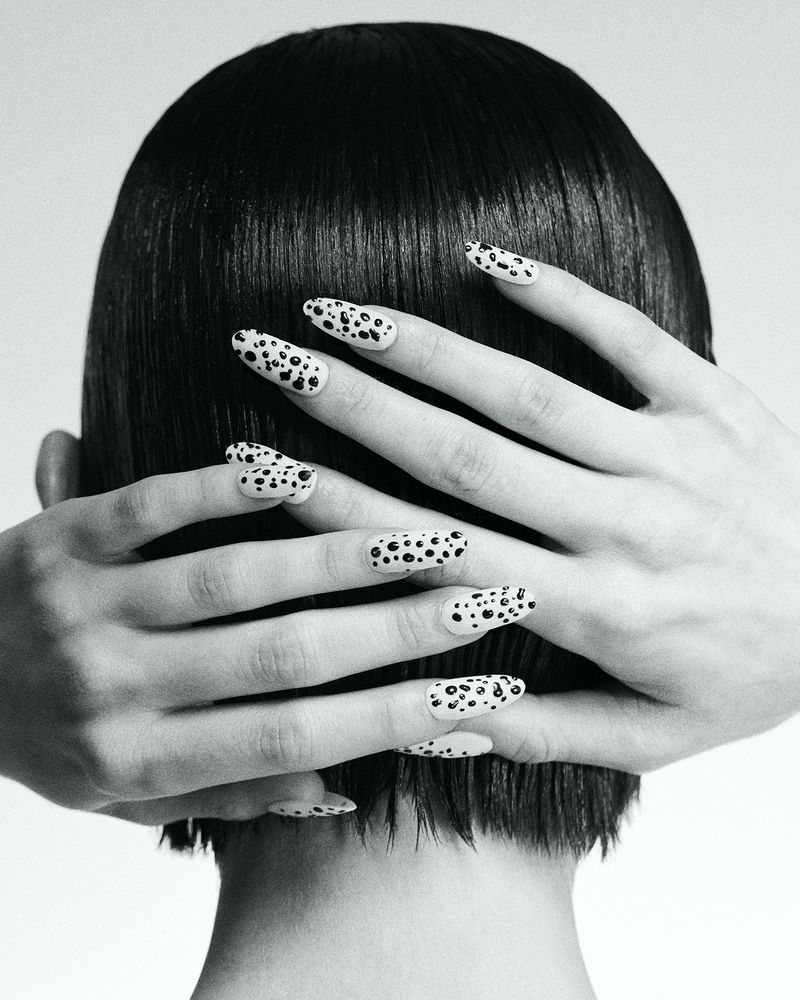 Back of a head with sleek dark short hair, featuring two hands crossed behind displaying black-spotted white manicured nails.