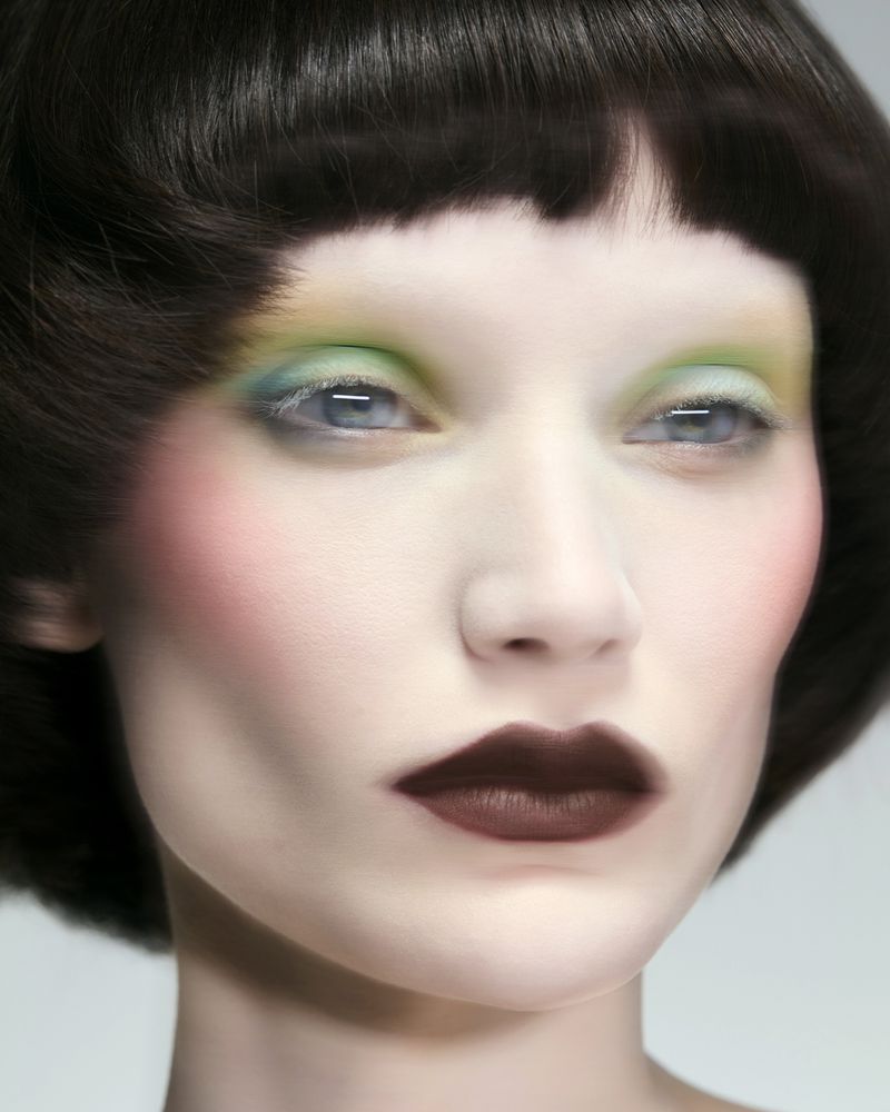 Close-up beauty portrait of a woman with short dark hair, gradient pastel eye makeup, pink blush, and dark matte lipstick.