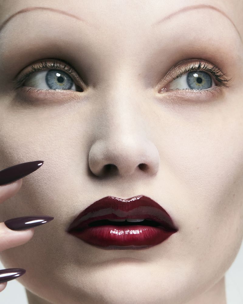 Close-up portrait of a model with thin drawn-on eyebrows, glossy burgundy lips, and long dark pointed nails near her face.