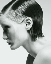 Side profile close-up portrait of a woman with slicked-back dark hair and graphic white makeup against a pale studio backdrop.