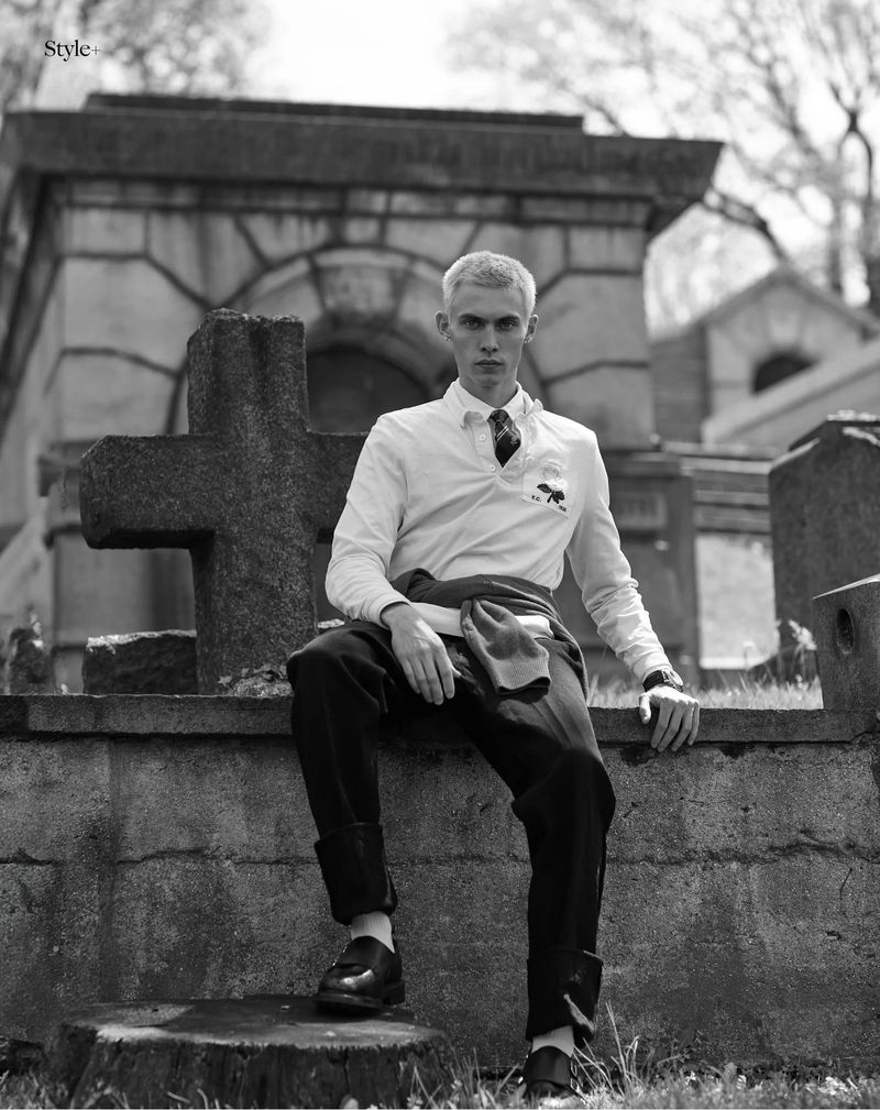 Model Arnau Sarda in a white collared shirt and black trousers sitting on a stone wall next to a cross in a cemetery.