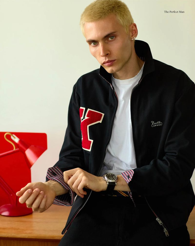 Blonde male model wearing a black zip jacket with red patch and adjusting cuffs beside a red desk lamp.