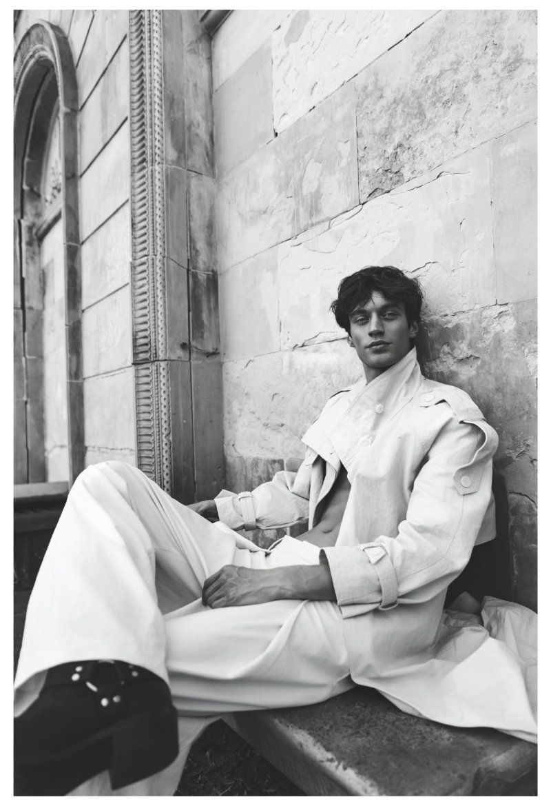 Model Piotr Kubiak seated outdoors wearing an open light trench coat, wide-leg trousers, and black platform boots against a stone wall.