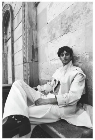 Model Piotr Kubiak seated outdoors wearing an open light trench coat, wide-leg trousers, and black platform boots against a stone wall.