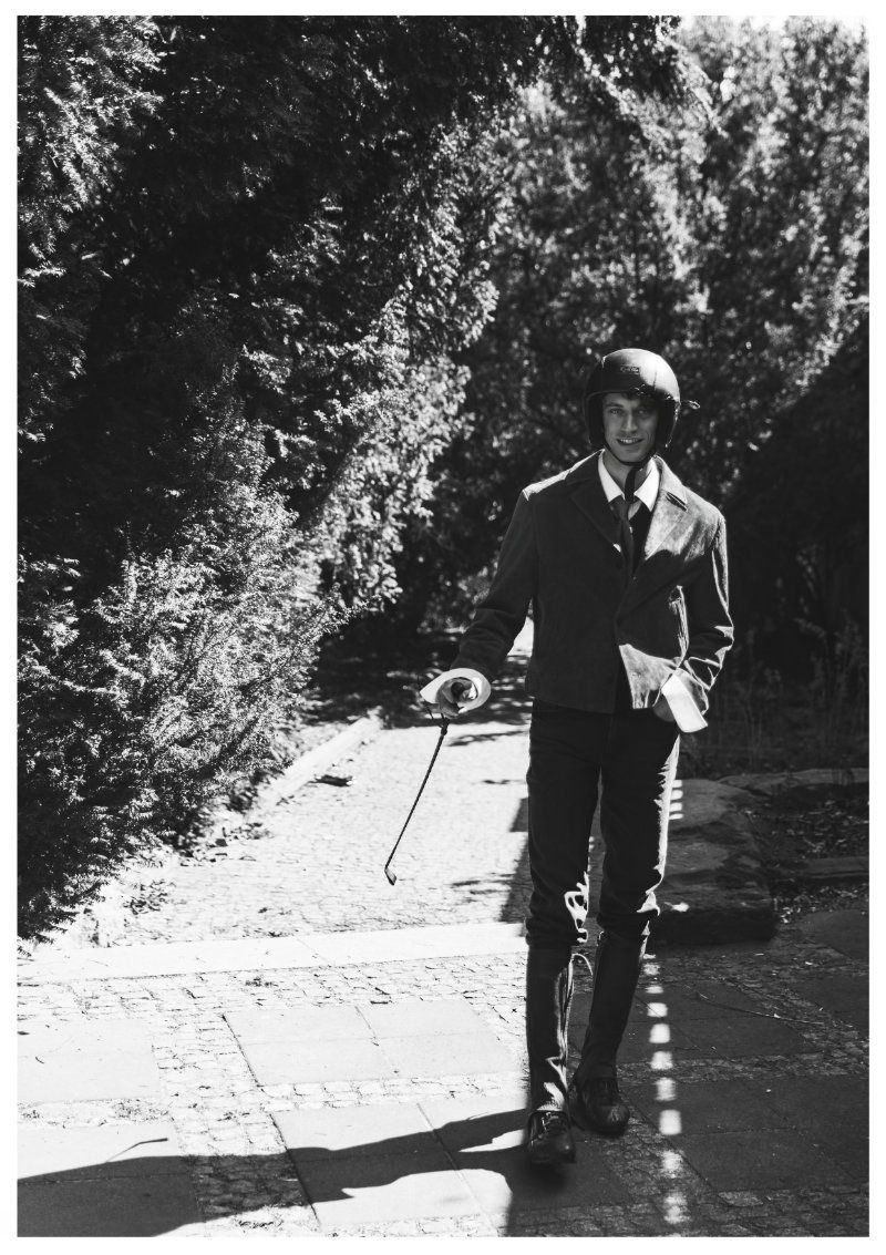 Model Piotr Kubiak in an equestrian helmet and blazer walking down a garden path holding a riding crop in a black and white photo.