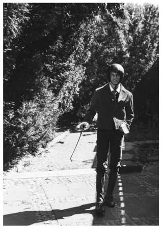 Model Piotr Kubiak in an equestrian helmet and blazer walking down a garden path holding a riding crop in a black and white photo.