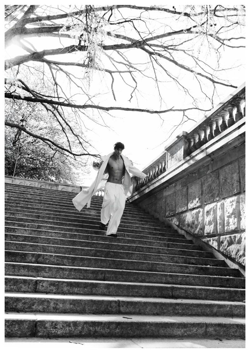 Model Piotr Kubiak in an open light coat and wide-leg white trousers walking down an outdoor stone staircase.