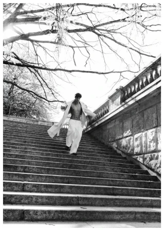 Model Piotr Kubiak in an open light coat and wide-leg white trousers walking down an outdoor stone staircase.