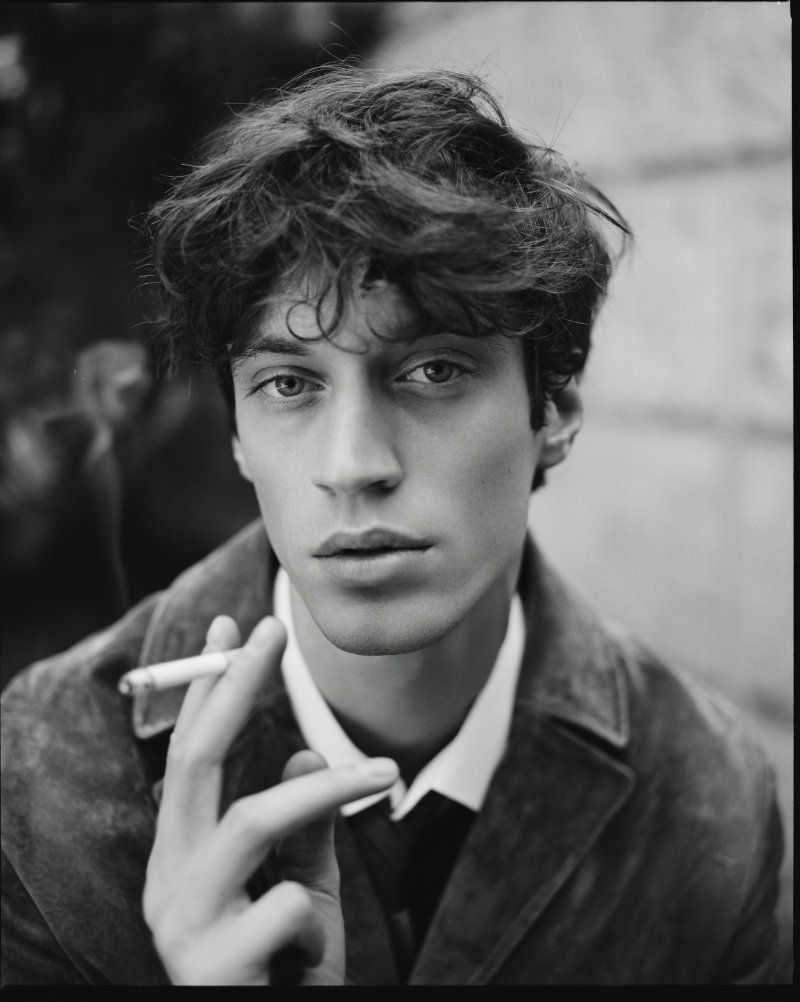 Black and white portrait of a man in a dark jacket, white collared shirt, and tie, holding a cigarette close to his face.