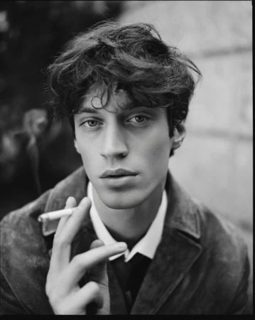 Black and white portrait of a man in a dark jacket, white collared shirt, and tie, holding a cigarette close to his face.