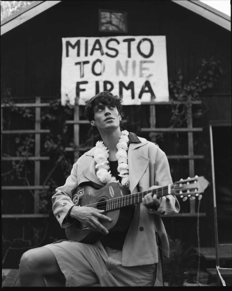 Young man wearing a light jacket, shorts, and a flower garland playing an acoustic guitar outside a dark wooden building.