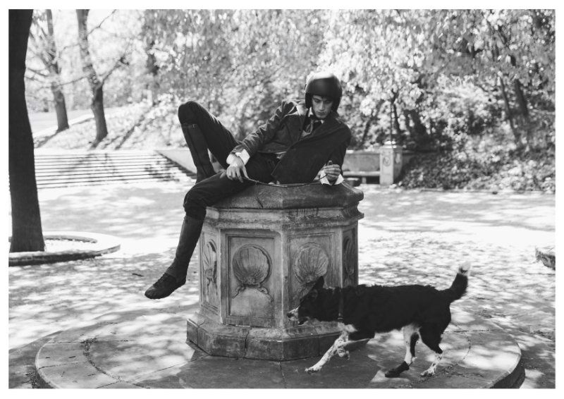 A man in an equestrian helmet and blazer lounges on a stone fountain pedestal outdoors next to a dog in black and white.
