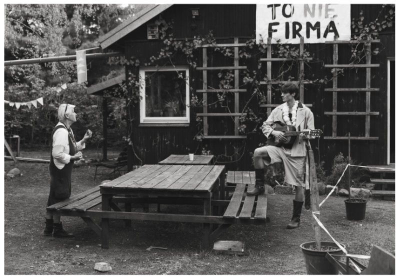 A man holding a guitar stands on a picnic bench outdoors opposite a woman outside a rustic dark wooden cabin.
