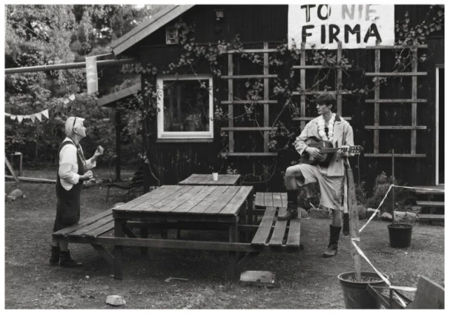 A man holding a guitar stands on a picnic bench outdoors opposite a woman outside a rustic dark wooden cabin.