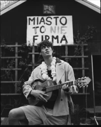 Young man wearing a light jacket, shorts, and a flower garland playing an acoustic guitar outside a dark wooden building.